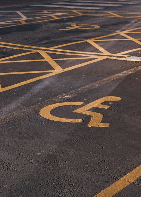 A close-up view of a designated accessible parking space on an asphalt surface, featuring prominent yellow wheelchair symbols painted on the ground. The markings include diagonal hatching lines and the international symbol for disability parking, situated adjacent to a building entrance or loading zone. The area appears clean and well-maintained, with additional parking lines and symbols visible in the background. This image emphasizes the clear identification of accessible parking spaces, which are often utilized during house removals or furniture transport to facilitate easy loading and unloading, aligning with the services provided by Man with Van Enfield in the context of safe home relocation and parking permits near Forty Hall.
