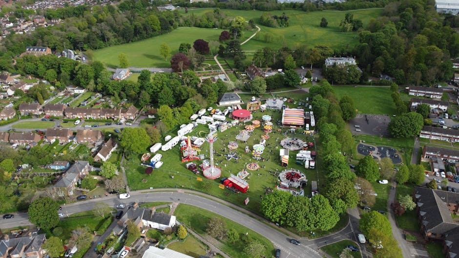 Aerial view of a fairground in a suburban area surrounded by residential houses, trees, and green fields, with a prominent loading area where Man with Van Enfield is involved in home relocation activities. The image shows numerous white and colorful vehicles, including vans and trailers, parked on the grass and pavement around the fairground. Several large cardboard boxes and furnished items are organized near trucks and vans, indicating packing and furniture transport during the loading process. Equipment such as trolleys, straps, and moving blankets are visible, suggesting an organized furniture transport operation as part of house removals. The fairground features amusement rides and tents, set amidst lush greenery, with a curved road bordering the site. The scene is illuminated by natural daylight, highlighting the logistics involved in a residential move, fitting for content about safe removals and parking permits in the Forty Hall area, ENFIELD, serviced by Man with Van Enfield.