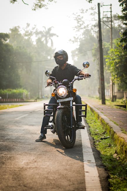 A person wearing a black helmet, mask, and dark clothing is seated on a black motorcycle parked on a paved road. The motorcycle is facing directly towards the camera with its headlight illuminated. The rider's feet are positioned on the ground, and they are holding the handlebars. Behind them, the road extends into a misty, tree-lined landscape with background utility poles and wires, suggesting a rural or suburban area. The scene appears to be during early morning or late afternoon with soft natural lighting. Although the image does not show furniture or household items, a professional home relocation or furniture transport company like Man with Van Enfield could be associated with efficiently managing moving logistics in nearby neighborhoods or narrow streets, particularly where careful planning of vehicle access is required.