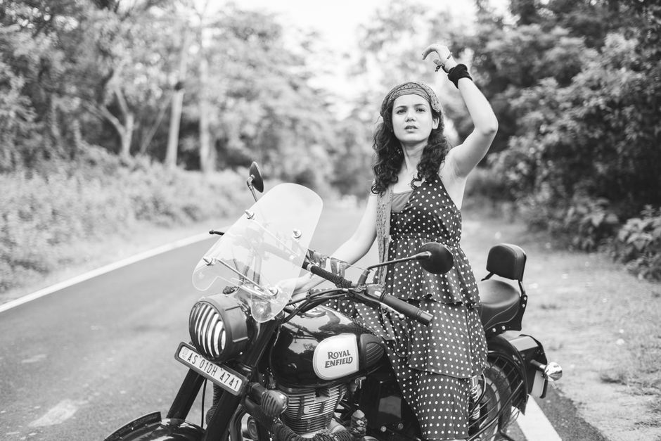 A woman with curly hair, wearing a polka dot dress and a headband, standing next to a Royal Enfield motorcycle on a quiet outdoor road, with trees and foliage in the background. She is lifting her left arm, holding a piece of clothing or fabric, while her right hand rests on the motorcycle handlebar. The motorcycle is equipped with a transparent windshield, side mirrors, and a black leather seat, suggesting it may be used for home relocation or transport of personal belongings. The scene appears to be set during daytime with natural light, and the environment is calm and rural. The setting reflects a moment in the packing and moving process, potentially related to furniture transport or home removals, as sometimes seen during logistics for house moves or local relocation services.