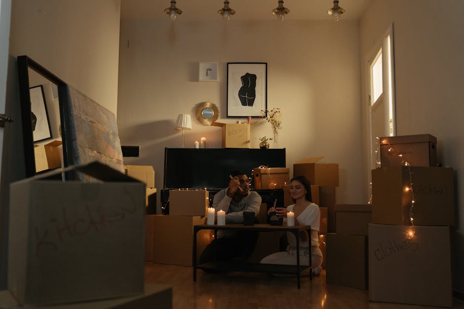 Inside a living room with beige walls and warm ambient lighting, a man with dark hair and a woman with long dark hair are seated on the floor among numerous cardboard boxes, some sealed with tape and marked with handwritten labels. The boxes vary in size and are stacked around them, with a few placed on a small coffee table also made of cardboard. The room features a black sofa behind the pair, adorned with candles and decorative items, and a flat-screen television on a low stand. Above the TV, a framed black and white line art print of a female silhouette hangs on the wall, flanked by a small lamp on the left and a vase with dried flowers on the right. To the right, a window allows natural light into the space. This scene captures the process of home packing and furniture transport preparation, as part of a house removal or relocation service by Man with Van Enfield.