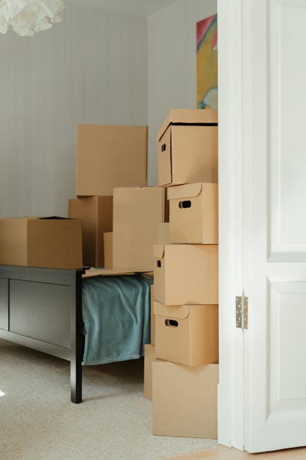 Inside a residential room during a home relocation, several brown cardboard boxes of various sizes are stacked on top of a grey wooden sideboard and directly on the floor, some partially covered with a light blue moving blanket. The boxes are sealed with clear packing tape and have integrated hand holes for easier handling. Natural light illuminates the room, revealing a partially open white door on the right, leading to a corridor or another room. A piece of abstract artwork or colourful painting is visible on the wall in the background. The scene depicts the packing stage of a furniture transport process, with boxes prepared for removal, supported by professional moving services such as Man with Van Enfield, who specialise in house removals and furniture transport, ensuring a smooth and organized home relocation.