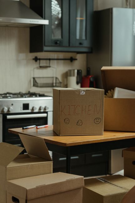 Inside a living room with beige walls and warm ambient lighting, a man with dark hair and a woman with long dark hair are seated on the floor among numerous cardboard boxes, some sealed with tape and marked with handwritten labels. The boxes vary in size and are stacked around them, with a few placed on a small coffee table also made of cardboard. The room features a black sofa behind the pair, adorned with candles and decorative items, and a flat-screen television on a low stand. Above the TV, a framed black and white line art print of a female silhouette hangs on the wall, flanked by a small lamp on the left and a vase with dried flowers on the right. To the right, a window allows natural light into the space. This scene captures the process of home packing and furniture transport preparation, as part of a house removal or relocation service by Man with Van Enfield.