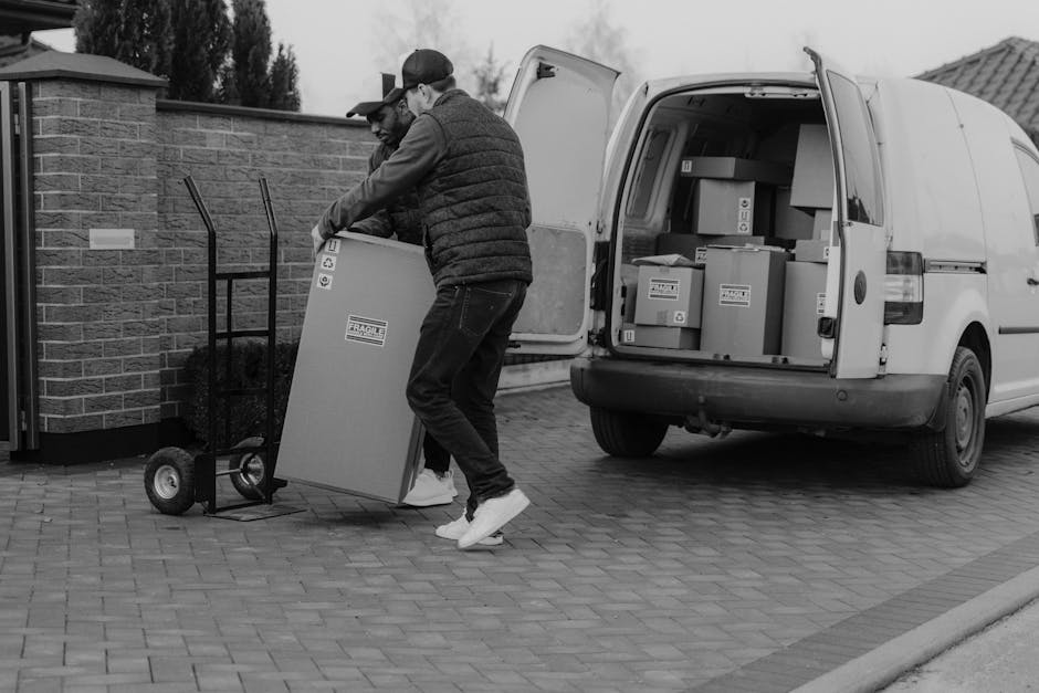 Two men are engaged in a home relocation process outdoors, loading cardboard boxes onto a trolley for transport, with some boxes placed inside the back of a white van parked on a brick driveway. One man is wearing a cap, and both are dressed in casual clothing, including jackets and sneakers. The open van reveals additional packed boxes arranged inside, ready for moving or delivery. Behind them, there is a brick wall and a building, with some trees and cloudy sky visible in the background. The scene captures a typical furniture transport and packing process during a removal service, with the men handling the boxes carefully, supported by equipment such as the trolley, as part of the loading process. The image reflects the logistical aspects of a house removal managed by [COMPANY_NAME], emphasizing professional moving activities characteristic of home relocation and packing and moving services.