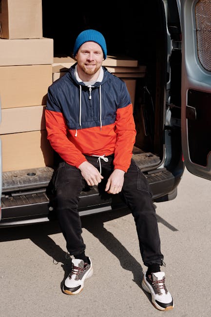 A man with a beard and light skin, dressed in a blue beanie, a navy and red jacket, black pants, and white sneakers, is sitting on the edge of an open van with its sliding door swung open. The interior of the van is visible, filled with neatly stacked cardboard boxes of various sizes, some wrapped in clear plastic for protection during the moving process. The boxes are organized along the side wall of the van, which is parked outdoors on a paved surface. The man appears relaxed and smiling, suggesting he is taking a break during a home relocation or furniture transport operation. The surrounding environment indicates a residential or urban area, with natural daylight illuminating the scene, capturing the logistics involved in a professional removals service, such as those offered by Man with Van Enfield.