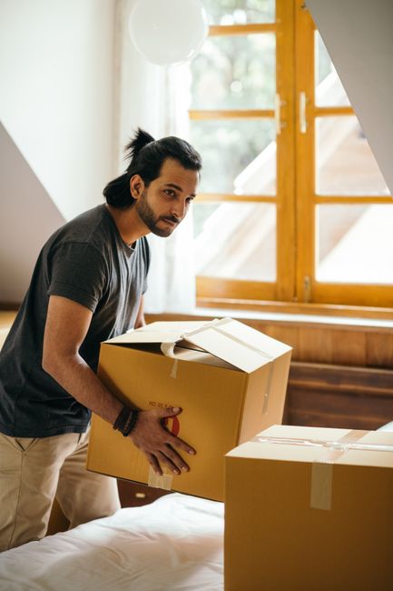 Two men are engaged in a home relocation process outdoors, loading cardboard boxes onto a trolley for transport, with some boxes placed inside the back of a white van parked on a brick driveway. One man is wearing a cap, and both are dressed in casual clothing, including jackets and sneakers. The open van reveals additional packed boxes arranged inside, ready for moving or delivery. Behind them, there is a brick wall and a building, with some trees and cloudy sky visible in the background. The scene captures a typical furniture transport and packing process during a removal service, with the men handling the boxes carefully, supported by equipment such as the trolley, as part of the loading process. The image reflects the logistical aspects of a house removal managed by [COMPANY_NAME], emphasizing professional moving activities characteristic of home relocation and packing and moving services.